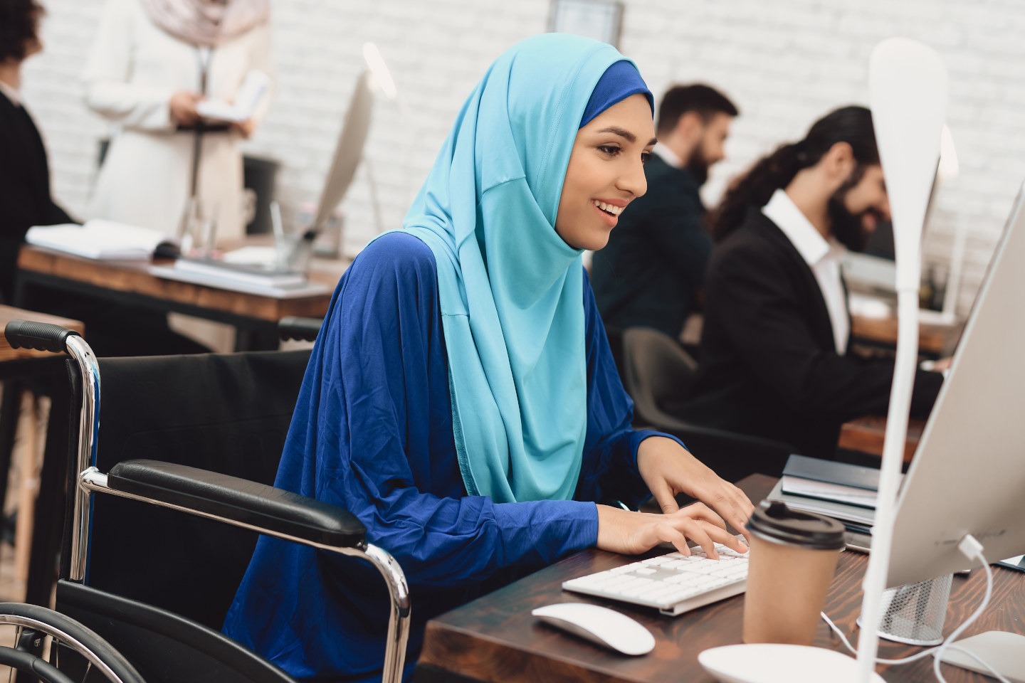 Disabled arab woman in hijab in wheelchair working in office. Woman is working on desktop computer.