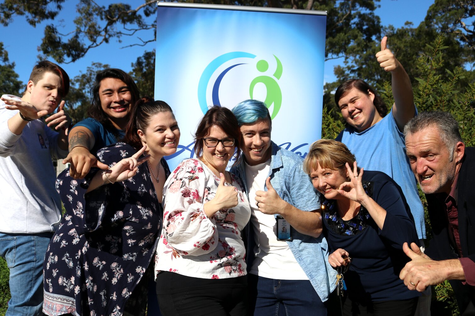 The image shows a group of people posing together outdoors in front of a banner. The banner features a blue gradient background with a green and blue logo, which appears to be a stylized human figure. The group members are making various hand gestures such as thumbs up and peace signs, and they seem to be in a celebratory or happy mood. The background includes trees and a clear sky, indicating that the photo was taken outside.

The purpose of the image is to capture a moment of camaraderie or celebration among the group members. This could be related to an event, gathering, or achievement that the group is celebrating. The cheerful and lively atmosphere suggests a positive and supportive environment, possibly highlighting the unity and shared joy of the group.