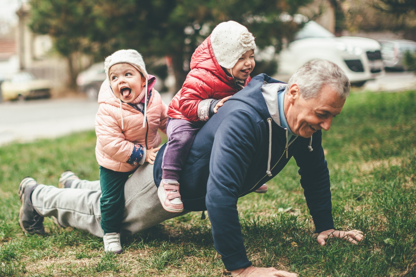 A photo of a playful grandfather and granddaughter. They are casually dressed and playing in the park. They exercise together. A grandfather is exercising while granddaughters are sitting on his back.