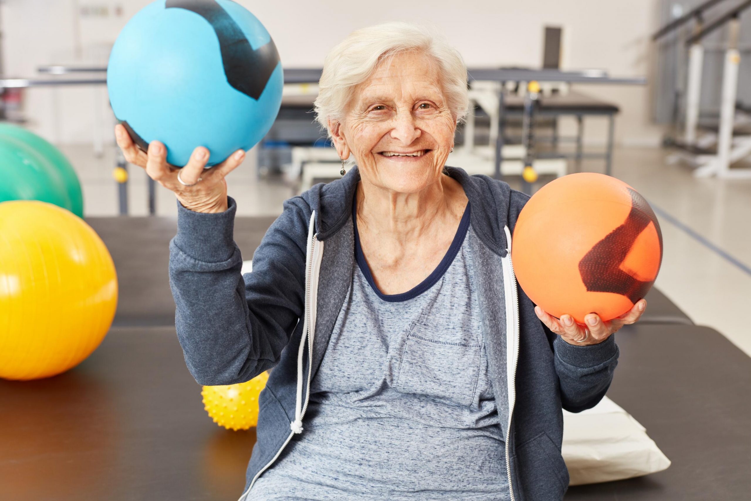 The image shows an older individual sitting and holding two balls, one in each hand. The ball in the left hand is blue with black markings, and the ball in the right hand is orange with black markings. The individual is wearing a grey shirt and a dark grey zip-up hoodie. In the background, there are other exercise balls, including a large yellow ball and a smaller yellow spiky ball. The setting appears to be a gym or physical therapy room, indicated by the presence of exercise equipment and the overall environment.

The purpose of this image is to highlight activities for maintaining or improving physical health and mobility, especially for older adults. The use of different types of balls suggests exercises that involve coordination, strength, and flexibility. This image is relevant as it showcases the importance of physical activity in promoting health and well-being, particularly in older age.