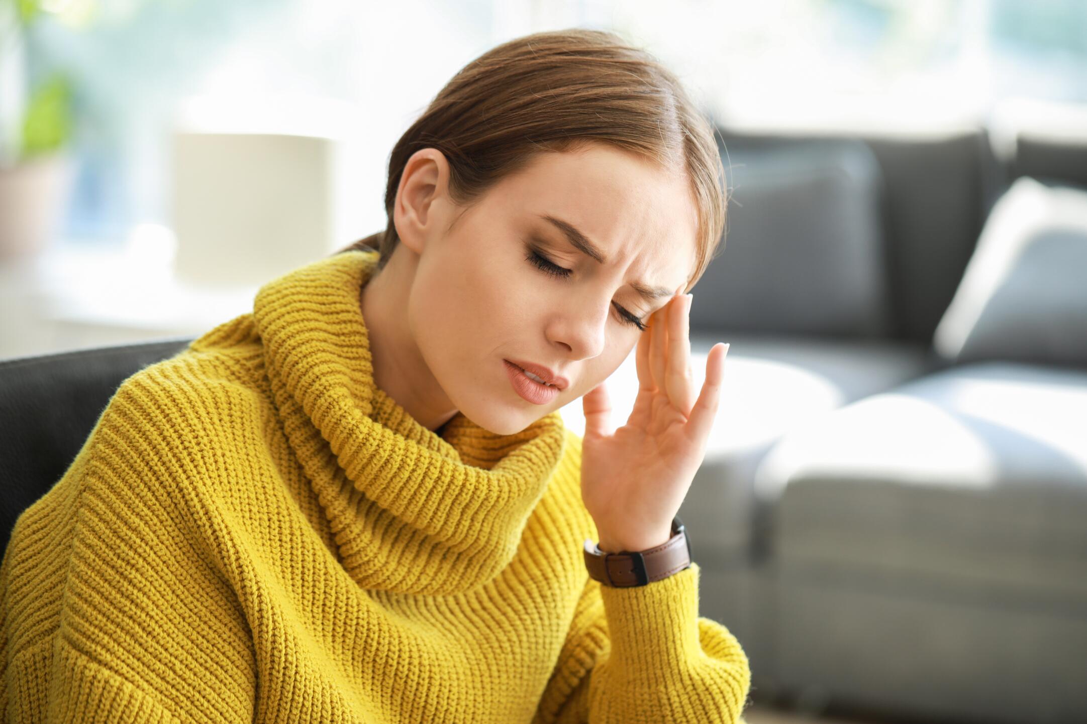 The image shows a person wearing a yellow knitted sweater with a high collar. They are holding a hand up to the side of their face. The background appears to be a living room with a couch and some blurred elements, suggesting a home environment. The yellow sweater stands out as a prominent feature in the image.

In a mental illness scenario, this image could depict someone experiencing a moment of anxiety or stress. The hand to the face gesture might indicate a coping mechanism or a moment of self-soothing. The cozy sweater and home setting could symbolize the importance of creating a comfortable and safe environment for oneself during challenging times. This image is used to raise awareness about mental health, emphasizing the need for self-care and the importance of seeking support when dealing with mental health issues.