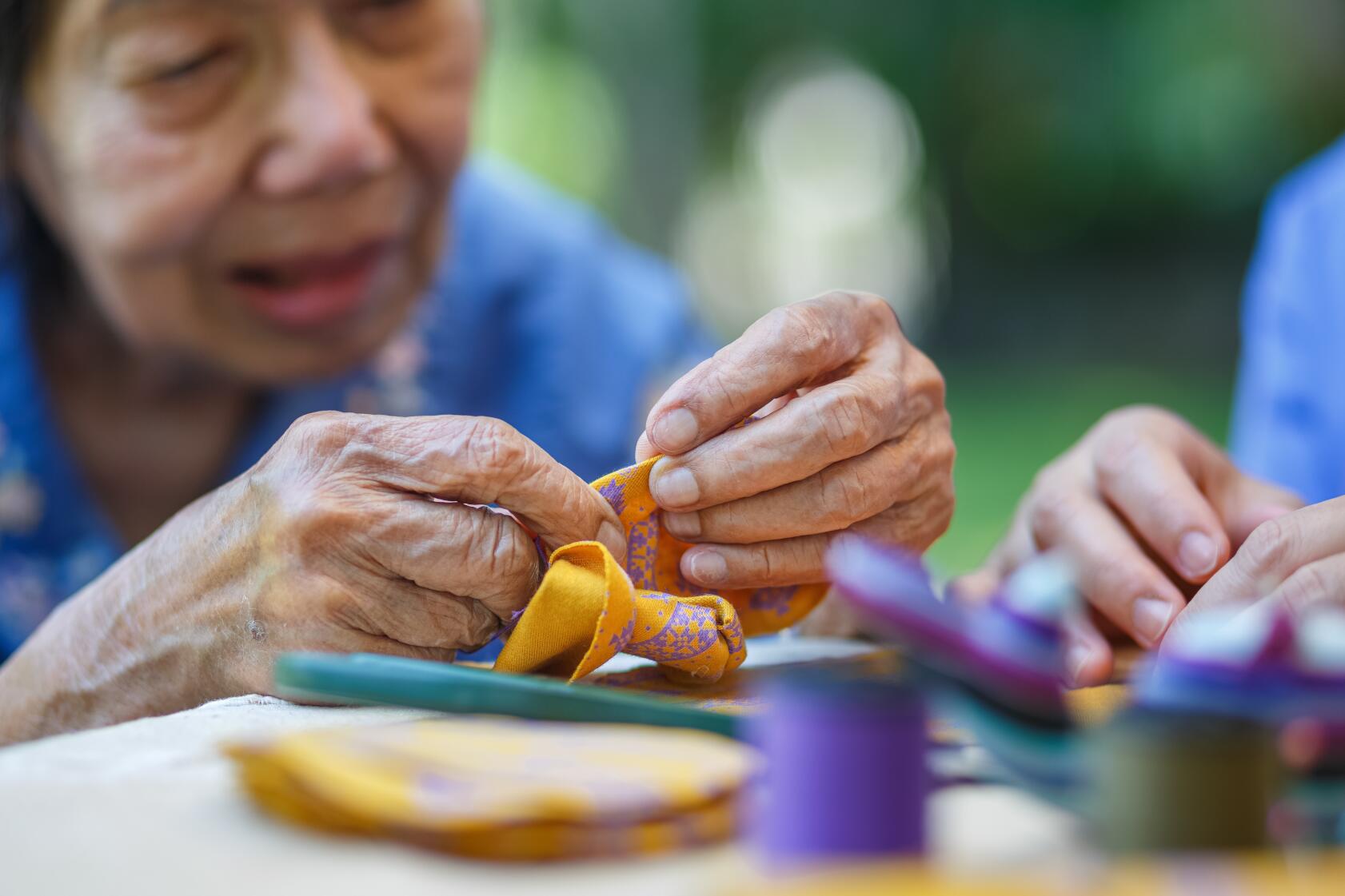 The image shows a close-up of two pairs of hands working on a piece of fabric. The hands appear to be engaged in a sewing or crafting activity. The fabric being worked on is yellow with a purple pattern. Various sewing tools and materials are visible in the foreground, including spools of thread and fabric pieces. The background is blurred, suggesting an outdoor setting.

The purpose of this image is to illustrate the process of crafting or sewing. It highlights the hands-on nature of such activities, which can be both therapeutic and engaging. The presence of multiple hands suggests a collaborative effort, possibly indicating a social or communal crafting session. This image is relevant for promoting the benefits of crafting, such as enhancing fine motor skills, fostering creativity, and encouraging social interaction.